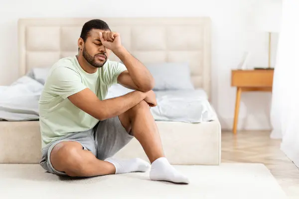 stock image Upset young black man in pajamas sitting on floor by bed at home, thinking about something, looking for solution while 2024 crisis, leaning on his hand, panorama with copy space