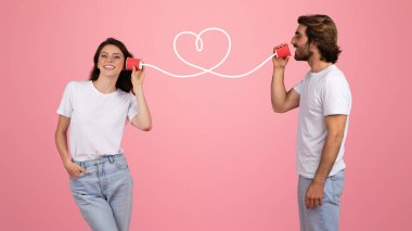 Kalp şeklinde bir telle konserve kutusuyla iletişim kuran gülümseyen Avrupalı kadın ve erkek, pembe arka plandaki beyaz tişörtlerle romantik bağlantıyı sembolize ediyor.