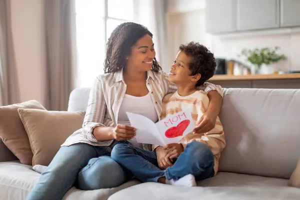 可爱的黑人儿子将红心手工制作的卡片送给他快乐的妈妈 在母亲们坐在客厅舒适的沙发上迎接母亲们的生日 复制空间 — 图库照片