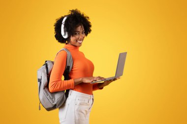 Kulaklıklı gülümseyen Afro-Amerikalı bir kadın, dizüstü bilgisayar kullanıyor, günlük turuncu bir balıkçı yaka giyiyor, omzunda gri bir sırt çantası var, sarı bir arka planda, stüdyoda.