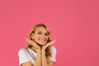 Yüzünde rüya gibi bir ifade ve neşeli bir gülümseme olan bin yıllık sarışın kadın terfi almak için boş bir alana bakıyor ve yüzünü pembe stüdyo arka planına yapıştırıyor. Güzellik ve moda sunar