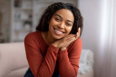 Güzel, kıvırcık saçlı, gülümseyen ve dinlenen bir kadın. Oturma odasında rahat bir kanepede dinlenirken. Afro-Amerikalı kadın kameraya poz veriyor.