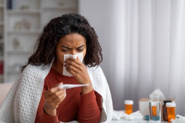 Hasta siyah kadın akan burnu temizliyor ve evde termometreye bakıyor, Afrika kökenli Amerikalı kadın oturma odasındaki kanepenin üzerinde battaniyeyle otururken sıcaklığı kontrol ediyor.