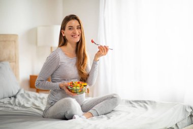 Sağlıklı Hamilelik Beslenme. Evde yatakta oturmuş taze sebze yiyen, lezzetli organik yiyeceklerle kaseyi tutan ve kameraya gülümseyen mutlu ve güzel hamile kadın.