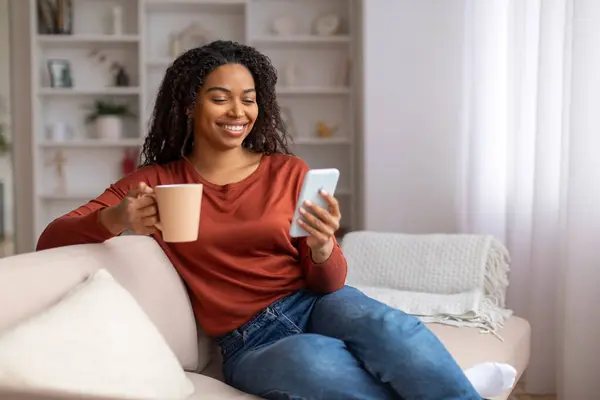 Young black woman relaxing on couch with coffee mug and smartphone, smiling african american female enjoying relaxed moment on comfortable sofa in cozy, well-decorated living room, copy space