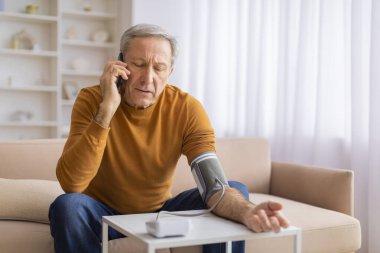 Endişeli yaşlı bir erkek kanepede oturuyor. Kan basıncı monitörü kullanıyor ve evinde telefonla konuşuyor.