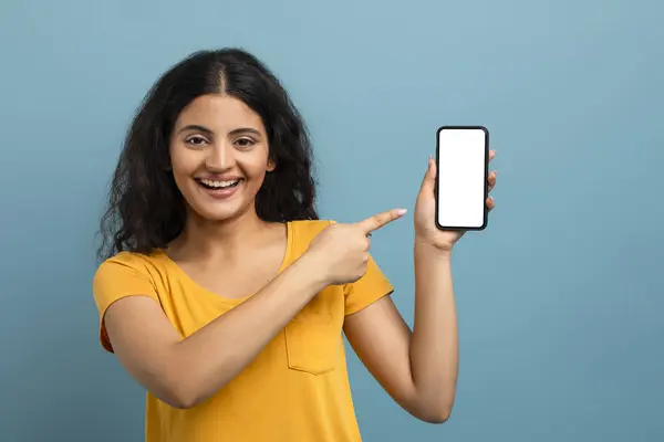 一个快乐的女人指着一个空白的智能手机屏幕 在沉闷的背景上带着大大的笑容 — 图库照片
