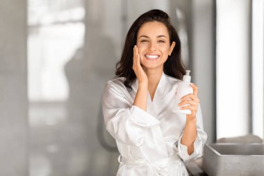 Beyaz ipeksi bornoz giyen neşeli, genç esmer kadın. Yanında da besleyici yüz kremi losyonu olan bir şişe taşıyor. Ortadoğulu güzel bayan banyoda nemlendirici cilt, yakın plan, kopyalama alanı
