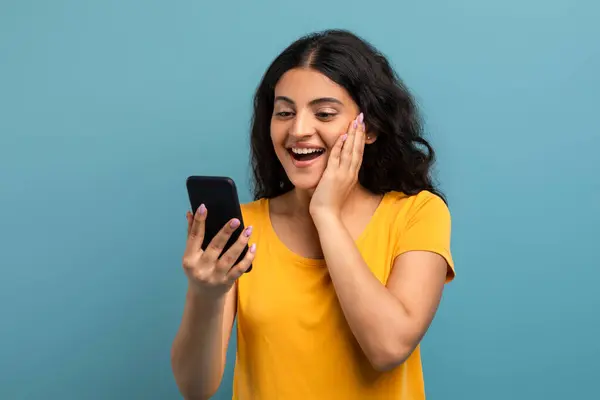一个穿着黄色T恤的兴奋女人正在看她的手机屏幕 带着惊讶的表情看着枯燥无味的背景 — 图库照片