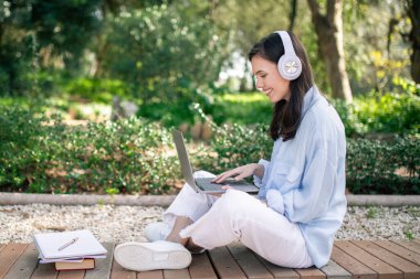 Huzurlu bir parkta dizüstü bilgisayar kullanan, yanında not defterleriyle gülümseyen Avrupalı genç bayan öğrenci, çalışırken ya da uzaktan çalışırken güzel bir günün tadını çıkarıyor.