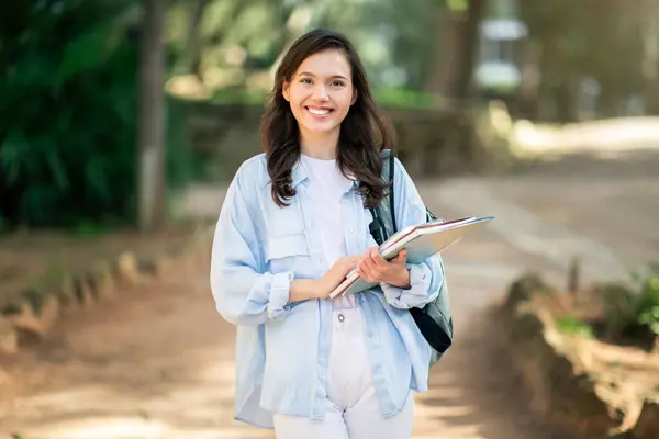 Happy european young woman student with a charming smile holding notebooks while confidently walking in a park, exemplifying a students life on campus, outdoor. Study, education