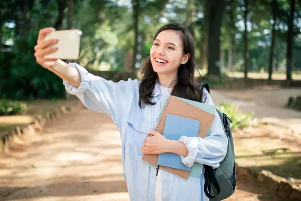 Exuberant european young woman student taking a selfie with her smartphone while holding textbooks in a verdant park, showcasing the joyful spirit of modern student life, outdoor
