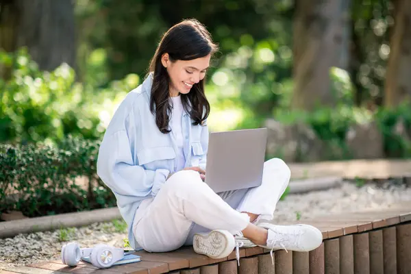 全神贯注的欧洲青年学生盘腿坐在郁郁葱葱的花园里的长椅上 在一个安静的室外环境中工作 旁边放着一台装有无线耳机的笔记本电脑 — 图库照片