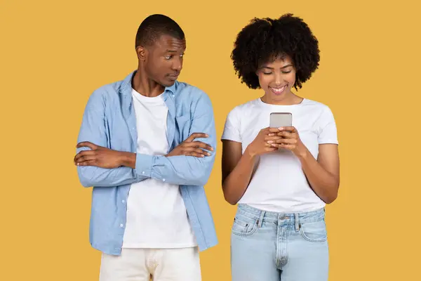 懐疑的なミレニアル世代のアフリカ系アメリカ人男性はスマートフォンに焦点を当てた笑顔の女性を見て 潜在的に黄色い背景での現代の関係における技術の問題を強調します — ストック写真