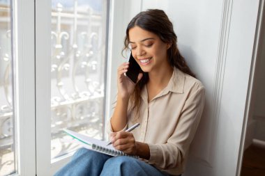 Neşeli genç bir kadın cep telefonuyla sohbet ediyor ve evde notlar alıyor, güzel bir milenyum kadını pencere kenarında rahat bir şekilde oturuyor, parlak bir iç mekanda birden fazla görev anı sergiliyor.