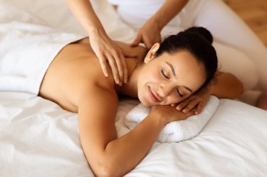 Modern spa ortamında rahatlatıcı sırt masajı sırasında gözleri kapalı uzanmış rahatlamış esmer bir kadın. Profesyonel masör müşterilere boyun ve omuz masajı yapıyor.