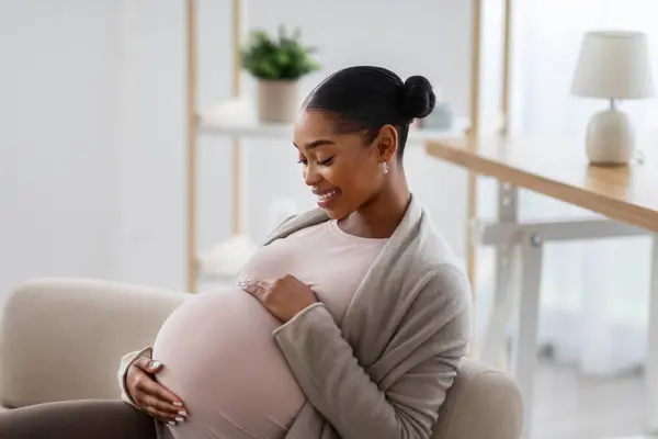 妊娠中の母親のコミュニケーション アフリカ系アメリカ人のサイドビュー 自宅でソファに座って彼女の大きな腹に触れ — ストック写真