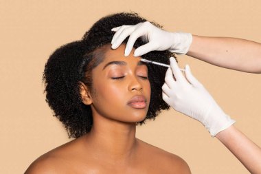 Serene black woman receiving meticulous eyebrow treatment, eyes closed in relaxation, as cosmetologist apply care with syringe on beige background clipart