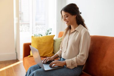 Evde koltukta otururken dizüstü bilgisayarla çalışırken gülümseyen ya da çalışan genç bir kadının portresi oturma odasında bilgisayarda yazı yazan, internette sörf yapan ya da e-posta yazan, boşluğu kopyalayan güzel bir kadın.