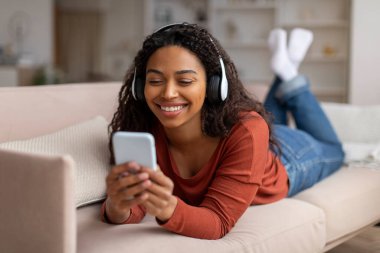 Gülümseyen siyah kadın portresi, kulaklık takan, akıllı telefonuyla evde dinlenen, neşeli, Afro-Amerikan bayan, oturma odasında dinlenirken cep telefonuyla müzik dinleyen.