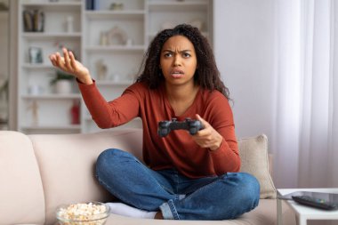 Oturma odasındaki koltukta oturan, elinde joystick ile strese girmiş, elinde video oyunlarında başarısız olan Afrikalı Amerikalı kadın, elinde jestler yaparak yakınıyor ve acı çekiyor.