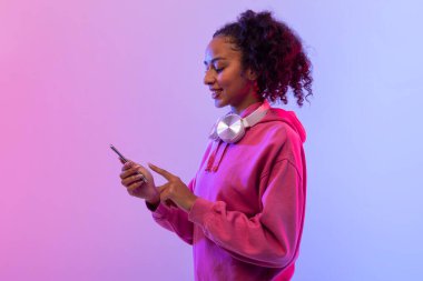 Kıvırcık saçlı, pembe kapüşonlu ve boynunda kulaklık olan mutlu siyahi kadın, akıllı telefonunu mor ve pembe gradyan arka planda kullanmaktan hoşlanıyor.