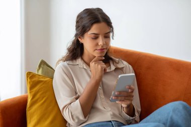 Düşünceli genç bir kadın, evinde akıllı telefondan mesaj okurken çenesine dokunuyor. Elinde cep telefonu olan, oturma odasındaki koltukta oturan, yakından çekilmiş bir fotoğraf.