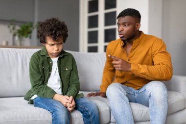 Bir baba oğluna ciddi bir konuşma yapıyor gibi görünüyor. Çocuk aşağı bakıyor ve babası onu işaret ediyor.