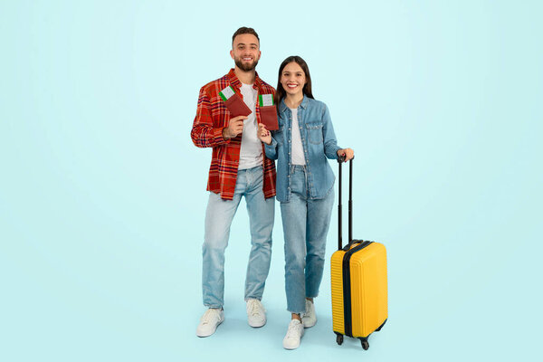 Cheerful caucasian travelers couple excitedly waiting for dream vacation, standing with travel tickets and suitcase, smiling as they plan romantic getaway, posing over blue backdrop. Full length