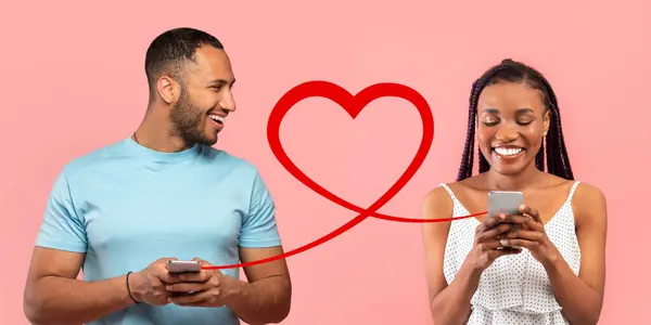 一个穿着蓝色T恤的迷人男人看着一个穿着白色圆点裙的快乐女人 两人都用红心图案的智能手机 这意味着他们之间的联系 背景是粉色的 — 图库照片