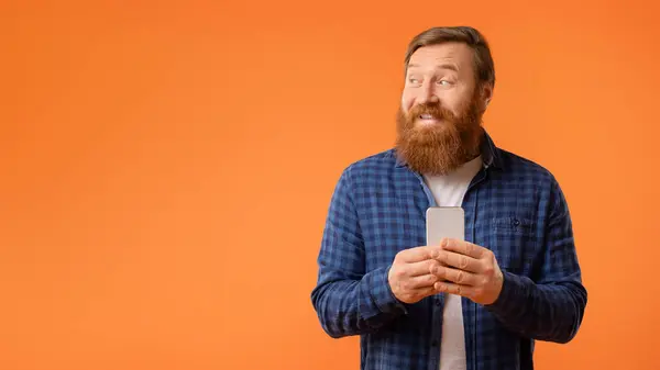 满脸笑容的红头发男人拿着手机 一边看空白处 一边嘲笑广告提供 一边发短信和在设备上使用应用程序 背景是橙色的 — 图库照片