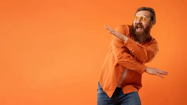 一头红头发的男人穿着鲜艳的橙色衬衫 戴着太阳镜 跳着滑稽的舞步 在工作室的背景上散发出凉爽的派对氛围和欢乐 有空白的文字空间 — 图库照片
