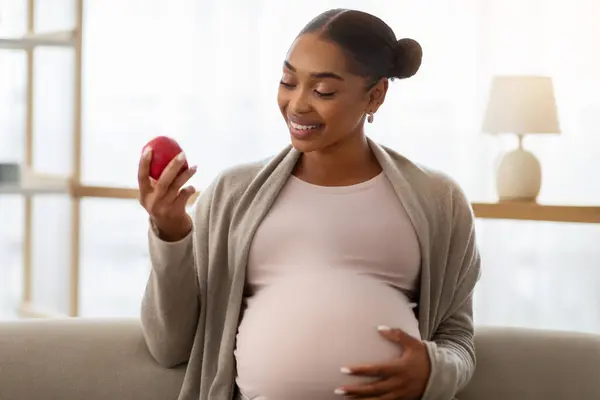 赤いリンゴを抱えている妊娠中のアフリカ系アメリカ人女性の肖像画は 自宅でソファーに座って 健康的なスナックを持っています 妊娠中のダイエット — ストック写真