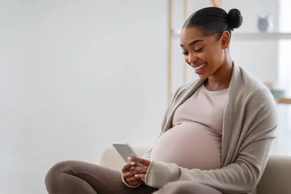 ポジティブな若い黒人妊娠中の女性は 自宅でソファーに座って 携帯電話を使用して 妊娠ブログを読んで 将来のお母さんのためのモバイルアプリを使用して スペースをコピー サイドビュー — ストック写真