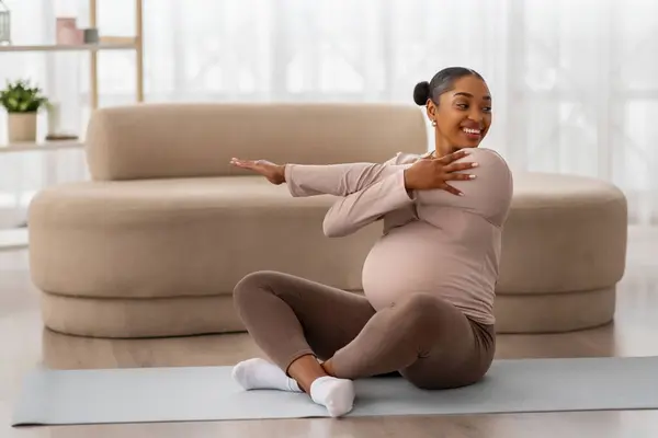 妊娠中のアフリカ系アメリカ人女性がリビングルームでヨガマットに座り 腕を伸ばし 空間を模倣しました ハッピーブラックは 家庭でスポーツをする女性を期待しています 妊娠中の健康的なライフスタイル — ストック写真