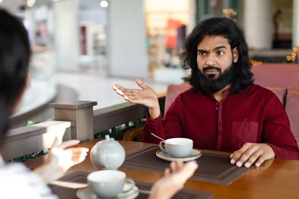 Confused bearded millennial indian man looking at his girlfriend and gesturing. Young eastern couple have date at cozy coffee shop, experiencing difficulties in relationships. Breakup, divorce