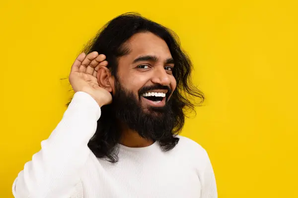 stock image Curious interested millennial indian man holding hand near ear and trying to hear information, yellow studio background, copy space. Excited eastern guy in casual clothing eavesdropping