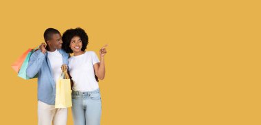 Binlerce yıllık Afro-Amerikan genç çifti ellerinde renkli alışveriş çantaları ve geniş sarı bir arka planda fotokopi alanına işaret ederek neşeli bir alışveriş deneyimi öneriyorlar.