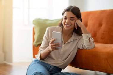 Gülümseyen, akıllı telefonlu, evinde yerde dinlenen, neşeli Avrupalı güzel bir kadının cep telefonuyla internete bakması ya da oturma odasında dinlenirken internetten alışveriş yapması.