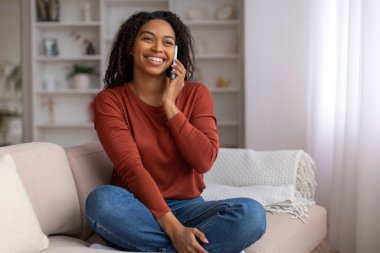 Güzel siyah bir kadın cep telefonuyla konuşuyor, gülümsüyor, Afro-Amerikan bir kadın cep telefonu konuşmasının tadını çıkarıyor, şık dekore edilmiş oturma odasında kanepede oturuyor.