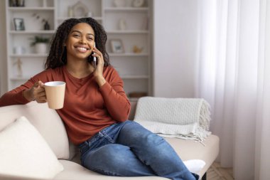 Neşeli genç siyah kadın cep telefonu sohbetinden ve evde kahve içmekten zevk alıyor. Afro-Amerikan kadın cep telefonuyla konuşuyor. Oturma odasında rahat bir şekilde koltukta oturuyor.