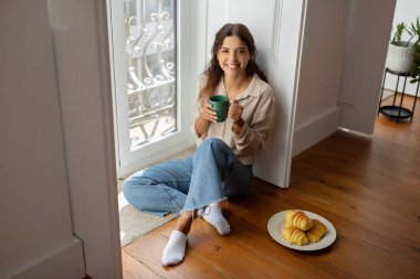Neşeli genç bir kadın evdeki pencerenin yanında dinlenirken kahvenin tadını çıkarıyor. Yanında bir tabak kruvasanla yerde oturan güzel bir kadın.