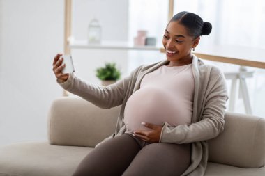 Neşeli afro-amerikan hamile kadın evde koltukta oturuyor, akıllı telefon kullanıyor, kocasıyla ya da arkadaşıyla internette görüşüyor, koca göbeğini gösteriyor.