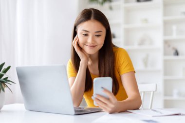 Akıllı telefonuna bakan gülümseyen Asyalı bir kadın ev ofisinde dizüstü bilgisayarla otururken güzel Koreli serbest çalışan bir kadın cep telefonundan mesaj okuyor veya video seyrediyor, uzay boşluğunu kopyalıyor.