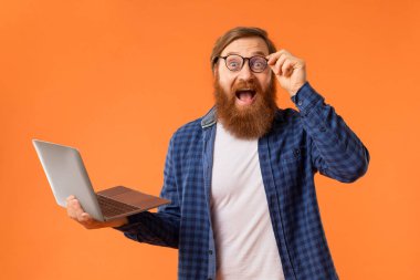 Heyecanlı kızıl saçlı sakallı programcı elinde dizüstü bilgisayarla kameraya sevinç ve heyecanla gülümsüyor turuncu stüdyo arka planında inanılmaz online teklife tepki veriyor.