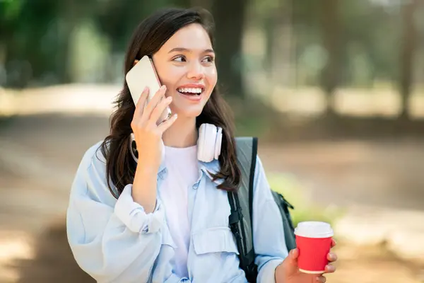 Animated european young woman student enjoying a phone conversation while casually holding a coffee cup and wearing headphones around her neck in a park setting, outdoor. Communication, gossip