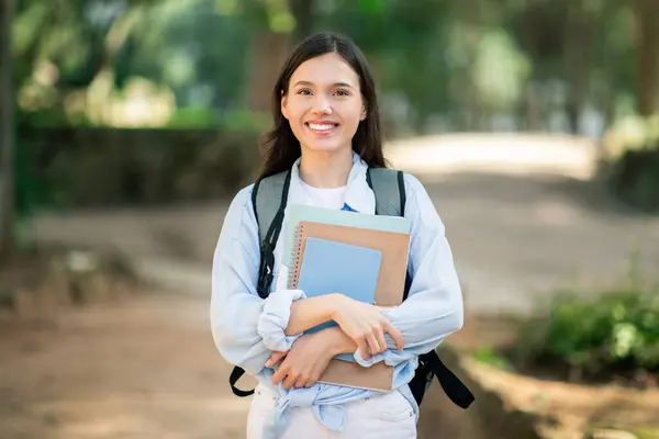 Cheerful european student college student lady with a radiant smile carrying textbooks and a backpack while walking through a lush green park on a sunny day, outdoor. Study, education