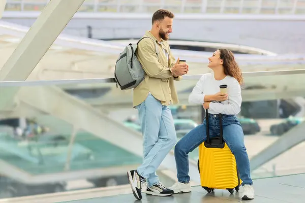 Happy Young Couple Having Fun While Waiting Flight At Airport, Cheerful European Man And Woman Drinking Takeaway Coffee And Chatting, Laughing And Enjoying Time Together, Lady Sitting On Suitcase