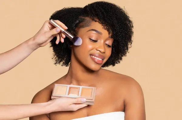 笑顔の若い黒人女性は 化粧アプリケーションの喜びを体現する彼女の頬骨 パレットを保持するアーティストに磨かれたハイライトを持っている楽しんでいます — ストック写真