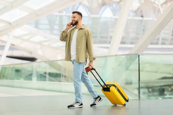 Handsome young man walking with suitcase at airport and talking on cellphone, smiling european guy carrying luggage while going to flight gate and enjoying mobile conversation, copy space